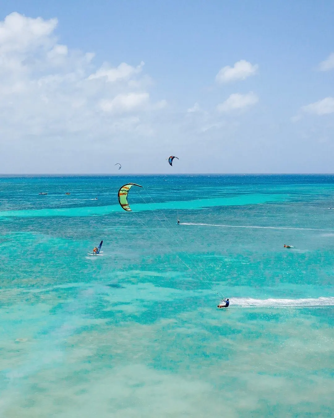 Vários kites e um windsurfista cruzando uma lagoa turquesa, vistos de cima