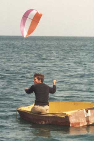 A man sits on the transom of a small yellow dinghy on open water, both arms raised and gripping short control lines, while a pink, white and orange arc-shaped kite flies low above the sea behind him.
