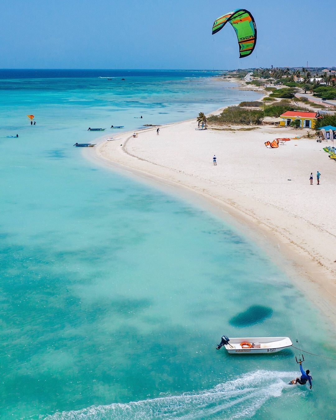 Aerial view of a kitesurfer carving along a Caribbean sandbar with a kite school on the beach